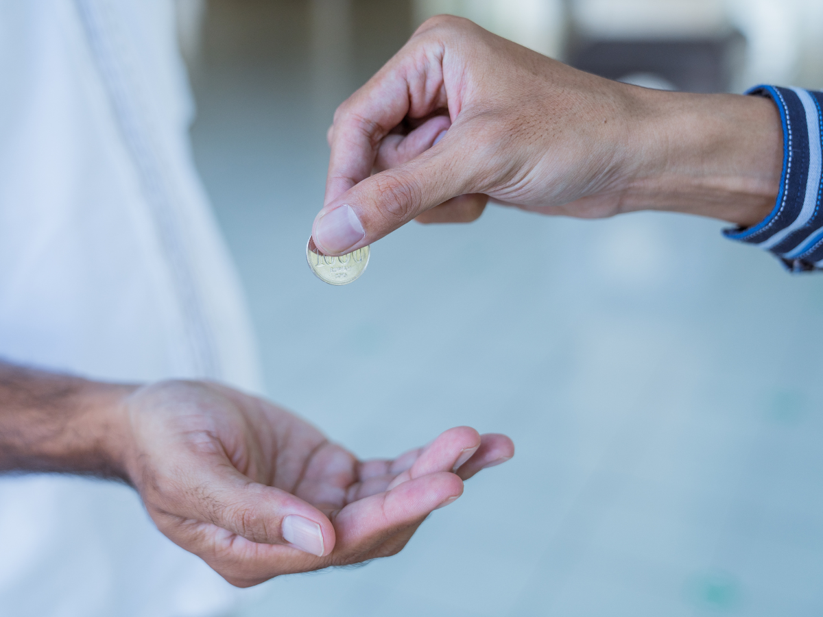 Two hands, one passing a coin to another, symbolizing the act of giving zakat or charity.
