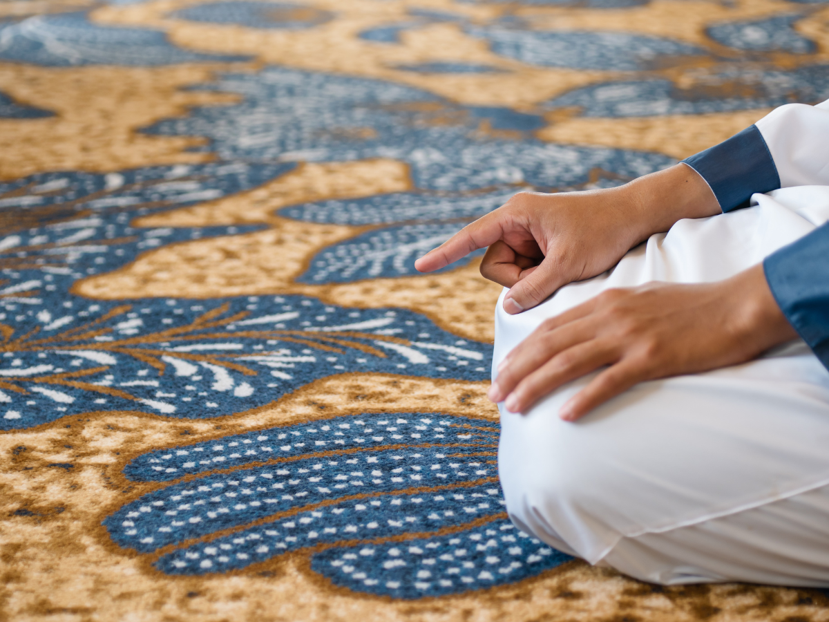 A person seated on a patterned prayer rug in a mosque, raising a finger during prayer, signifying a part of the Islamic prayer ritual.