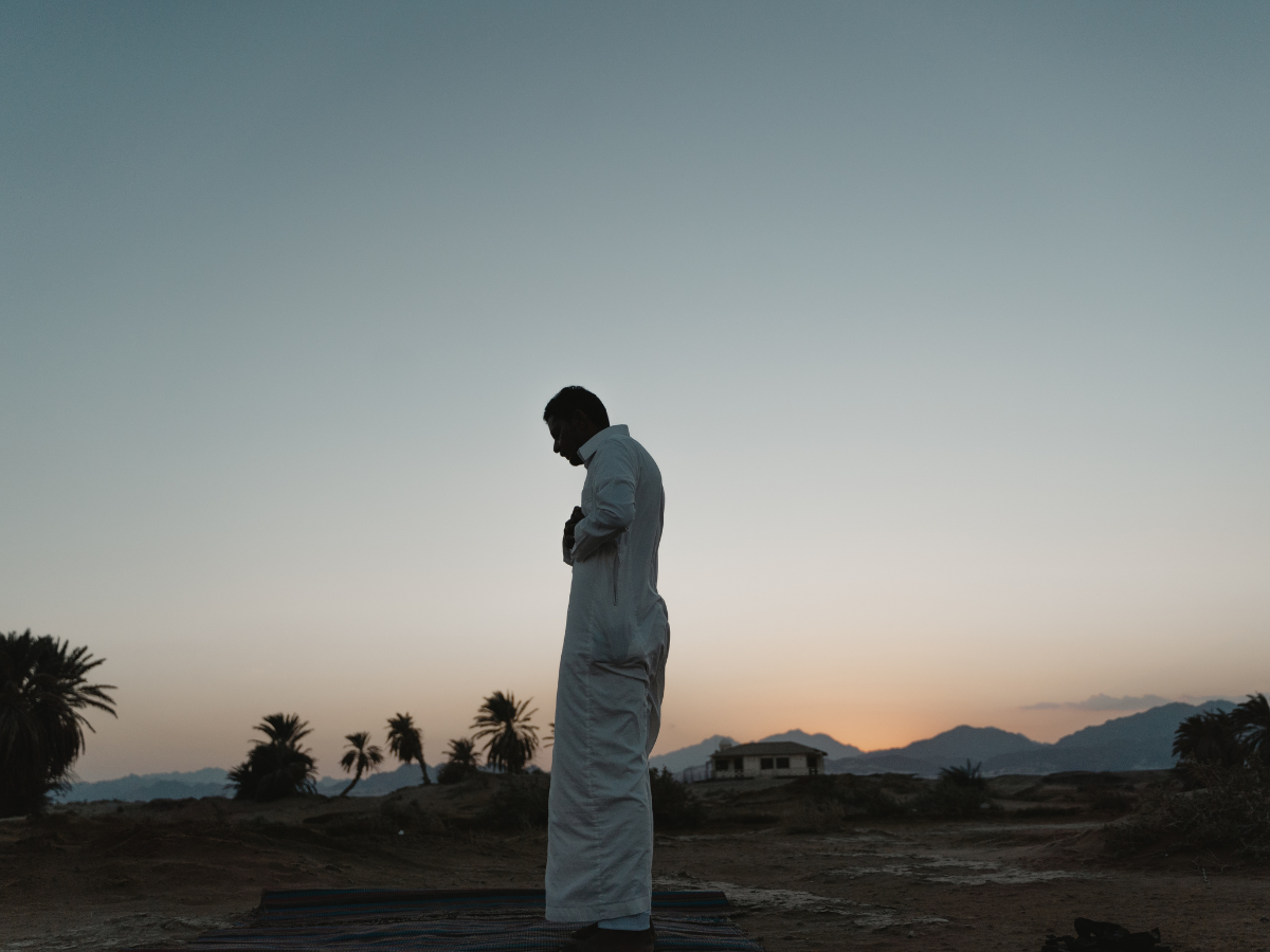 A man in traditional white attire praying outdoors at dusk, with palm trees and a dimly lit sky in the background